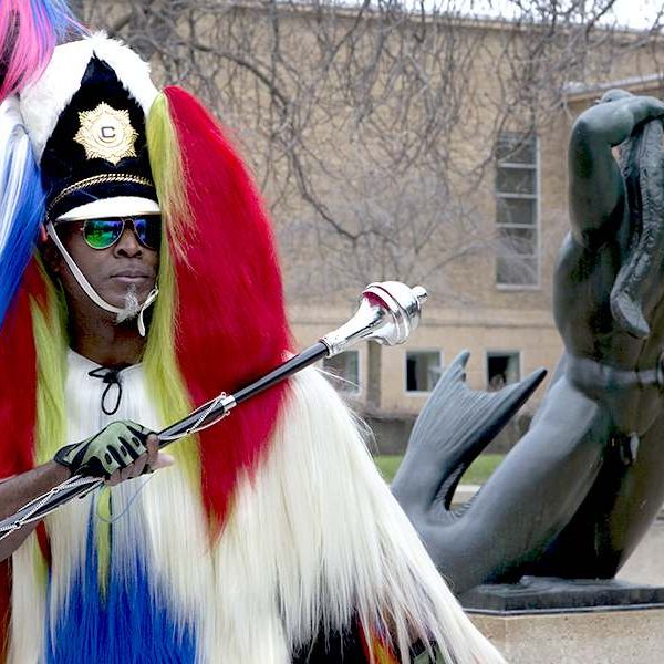 man dressed in colorful furs holder saber next to statue