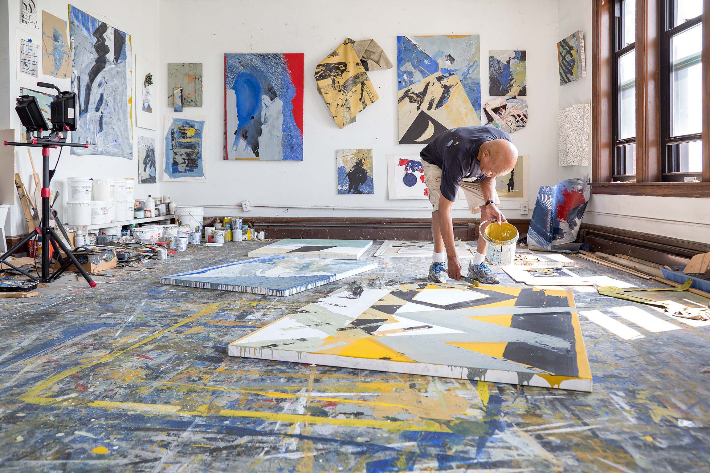 Person leaning over with brush in hand to apply paint to large canvas laying horizontally on the floor. The person is painting in a studio brightly lit with natural light from windows in the right of frame. Various paintings are hung around the walls. A large paint spattered tarp covers the floor.