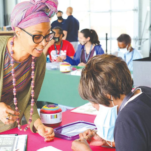 A person wearing a colorful hair wrap and necklace leans over to help someone with a facemask work on a craft project on a red table. In the background, more people are seated at another table also working on a pouch making project