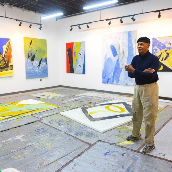 Artist Allie McGhee stands in a room with white walls. Several paintings are arranged on the two visible walls. A couple in progress paintings are in progress on the floor.